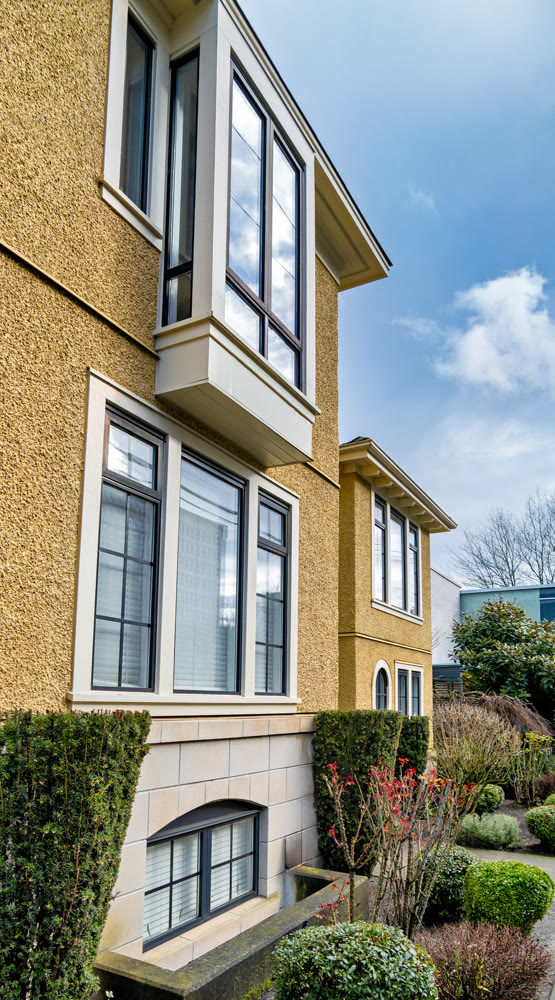 Yellow stucco house exterior with bay window and shrubs under a cloudy blue sky