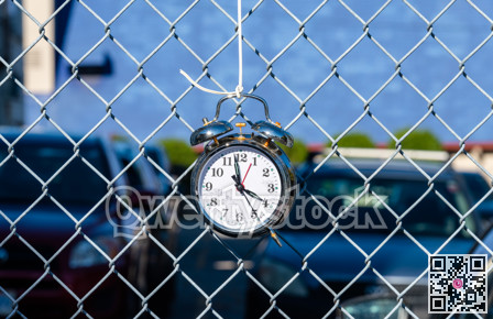 Old alarm clock hanging on metal net fence with cars parked on the backgound