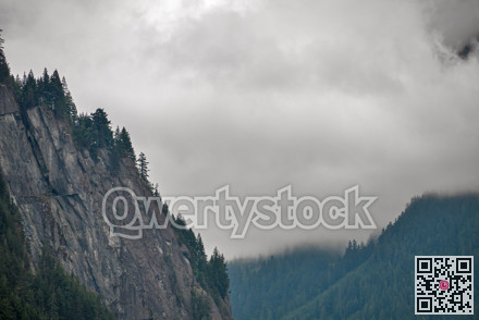 Steep rocky mountain cliff surrounded by trees on overcast sky background